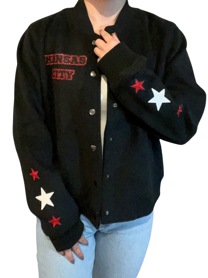 Puede incluir: Chaqueta bomber negra con parches de estrellas rojas y blancas en las mangas. La chaqueta tiene un parche rojo "Kansas City" en la parte delantera.
