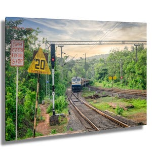 May include: A white and blue train travels on a track through a lush green forest. A yellow triangular sign with the number 20 is visible in the foreground. The sign reads "20 Kmph" in red letters. A red traffic light is visible in the background.