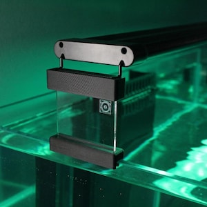 Op de afbeelding: Zwarte aquariumverlichtingsarmatuur gemonteerd boven een glazen tank gevuld met water. De armatuur heeft een rechthoekige vorm en is met zwarte beugels aan de tank bevestigd. Het water wordt verlicht met een groene gloed.