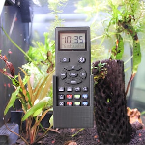 Op de afbeelding: Een zwarte aquariumcontroller met een digitaal display dat 22:35 uur weergeeft. Het heeft knoppen voor "Timer", "Clock" en "DIY", evenals verschillende pictogrammen. De controller bevindt zich voor een aquarium met waterplanten.