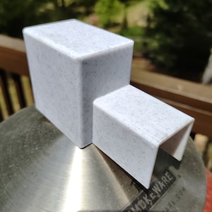 May include: White plastic adapter with a speckled finish. The adapter has a square top and a rectangular bottom with a slot opening. The adapter is sitting on a stainless steel surface with the text "Smokeware" and "Smokeware.com" printed on it.