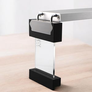 May include: A contemporary desk lamp with a rectangular silver metal arm and a black base. The lamp features a clear acrylic panel and a grey accent piece. The lamp is on a wooden surface.