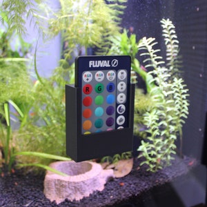 May include: A black plastic holder with a blue remote control for an aquarium light. The remote has a Fluval logo and buttons for white and RGB lighting.