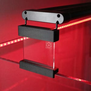 Puede incluir: Luminaria de acuario negra con un panel de vidrio transparente. La luminaria está montada en un panel de vidrio con una tira de luz LED roja. El panel de vidrio tiene un diseño circular grabado.