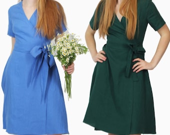 May include: Two women wearing wrap dresses, one in blue and one in green. Both dresses have a tie at the waist. The woman in the blue dress is holding a bouquet of white flowers.