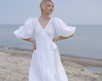 Könnte beinhalten: Ein weißes Leinen-Wickelkleid mit Puffärmeln. Das Kleid ist knielang und hat einen V-Ausschnitt. Das Model steht auf einem Sandstrand.