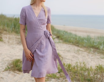 May include: A woman wearing a lavender linen wrap dress with short sleeves and a tie at the waist. She is walking on a sandy beach with green grass in the background.