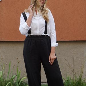 Op de afbeelding: Een wit overhemd met zwarte bretels, gecombineerd met een zwarte broek met wijde pijpen. De outfit wordt gecompleteerd met bruine sandalen. Het model staat voor een bruin en grijs gebouw.