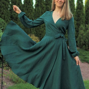 May include: A woman wearing a long-sleeved, emerald green wrap dress with a V-neckline and a tie at the waist. The dress has a flowing skirt and the woman is standing in a grassy area with trees in the background.