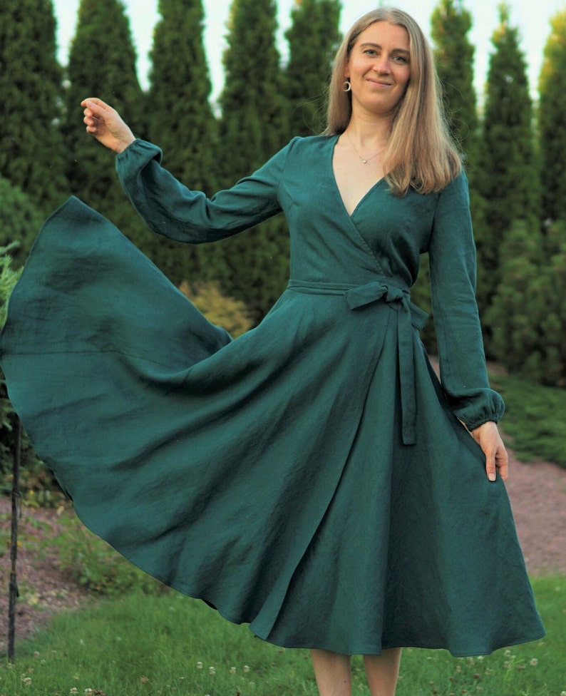 May include: A woman wearing a long-sleeved, emerald green wrap dress with a V-neckline and a tie at the waist. The dress has a flowing skirt and the woman is standing in a grassy area with trees in the background.