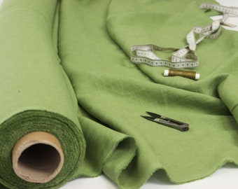 May include: A roll of light green fabric with a measuring tape, thread, and small scissors. The fabric is partially unrolled, showing its texture and color. The tools suggest sewing or crafting.