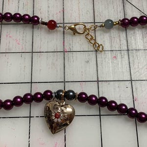 May include: A necklace featuring alternating dark gray and burgundy-colored beads. The necklace has a gold-colored clasp and a heart-shaped pendant with red accents. Additional charms include a gold flower and a key.