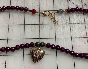 May include: A necklace featuring alternating dark gray and burgundy-colored beads. The necklace has a gold-colored clasp and a heart-shaped pendant with red accents. Additional charms include a gold flower and a key.