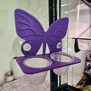 May include: Purple butterfly-shaped feeder with two clear plastic cups attached to a glass surface with suction cups.