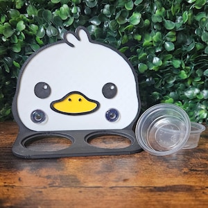 May include: A white and black duck-shaped sauce holder with a yellow beak and two clear plastic sauce cups. The holder has two circular openings and is set on a wooden surface with a green leafy background.
