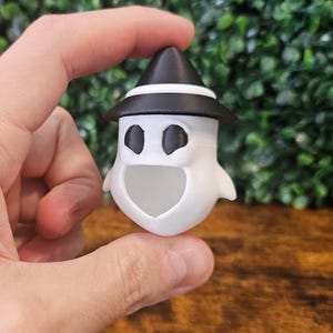 Könnte beinhalten: Eine weiße Geisterfigur mit einem schwarzen Hexenhut und einem schwarz-weiß gestreiften Band. Der Geist hat einen schwarzen Mund und zwei schwarze Augen.