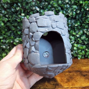 Peut inclure: Un pot de fleurs en forme de château en pierre grise avec une ouverture arrondie. Le pot est fait d'un matériau texturé qui ressemble à de la pierre.