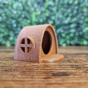 Op de afbeelding: Een klein, bruin, houten huis met een rond raam en een klein veranda. Het huis is gemaakt van een getextureerd materiaal en heeft een rustieke uitstraling.