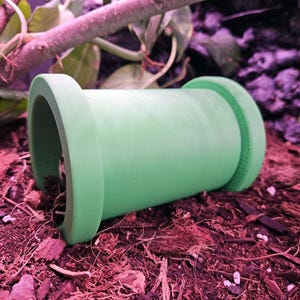 Puede incluir: Un tubo de plástico verde con una superficie lisa y un borde ligeramente texturizado. El tubo está sobre una cama de mantillo marrón.