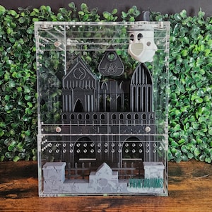May include: A clear acrylic display case featuring a black gothic castle design with a white ghost figure. The castle has detailed architectural elements. The bottom of the case has the text "PRINT AND WORKS".