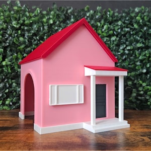 May include: A pink miniature house with a red roof, white trim, and a dark grey door. The house has an arched opening on the side and a small porch. The background is a green hedge.