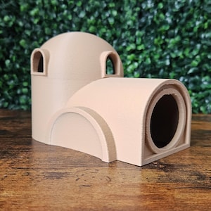 May include: A tan-coloured, 3D-printed structure with a domed top and arched openings. The design features a cylindrical tunnel and a curved base, resembling a miniature architectural model. The object is set against a backdrop of green foliage.
