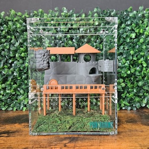 Puede incluir: Un terrario de acrílico transparente con una escena de castillo en miniatura en su interior. El castillo presenta muros grises, tejados naranjas y un puente de madera. Musgo verde y un pequeño acento azul en la base. El terrario está sobre un fondo de follaje verde.