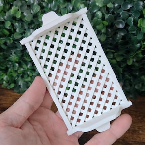 May include: A white, rectangular trellis with a lattice design. The trellis has a decorative top and bottom edge. The background features green foliage and a wooden surface.