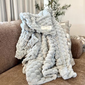 May include: A soft, light gray blanket with a textured, bubble-like pattern is draped over a brown sofa. The blanket has a plush appearance and a tag that reads "Bless & Boutique". The blanket is folded over a decorative pillow.