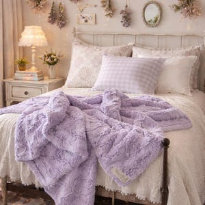 May include: A soft, lavender-colored faux fur throw blanket draped over a bed. The blanket has a textured, plush surface and is paired with white and lavender pillows and bedding. A nightstand with a lamp and books is visible.