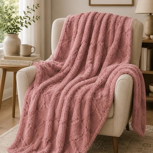 May include: A plush, dusty rose-colored throw blanket with a textured, raised diamond pattern. The fabric has a soft, fuzzy appearance and is draped over a light-colored upholstered armchair, showcasing its thick, warm, and cozy material.