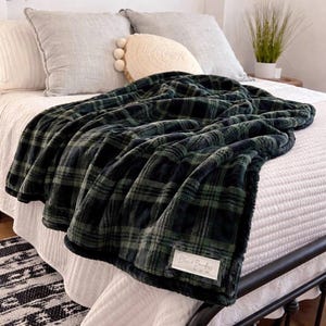 May include: A plush, dark green and black plaid blanket draped over a bed. The blanket has a soft, textured appearance and a label that reads "Bliss & Boundless". The bed is made with white linens and has gray pillows.
