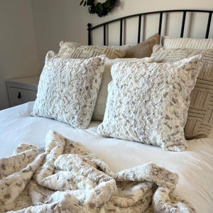 May include: A bed is made with white bedding, fluffy white and gray pillows, and a matching throw blanket. The pillows have a textured, fur-like appearance. The throw blanket is folded at the foot of the bed.