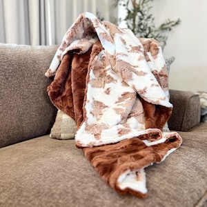 May include: A brown and white faux fur throw blanket with a soft, plush texture. The blanket is folded and draped over a brown sofa.
