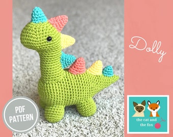 May include: A handmade green crochet dinosaur toy with colorful spikes in shades of blue, pink, and yellow. The toy has black safety eyes and is labeled "Dolly" in cursive. A "PDF Pattern" label is also visible.