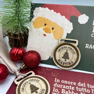 Può includere: Due ornamenti natalizi in legno con un disegno di albero e le parole "Orgoglioso di" e i nomi "Martina" e "Claudio". Gli ornamenti sono su uno sfondo rosso e verde con un'illustrazione di Babbo Natale.