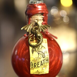 Puede incluir: Una botella redonda de vidrio rojo con un dije de dragón decorativo y una etiqueta que dice "Dragon's Breath". La botella está sellada con cera roja y tiene un tapón de vidrio transparente. La etiqueta también dice "Moonlight Apothecary".