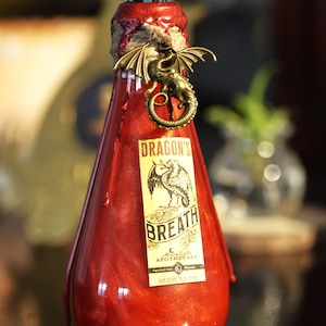 May include: A glass bottle filled with shimmering red liquid, sealed with red wax and twine. A metallic gold dragon charm is attached to the neck. The label reads DRAGON'S BREATH, Moonlight Apothecary, and READY TO USE. A blue glass bead sits atop the wax seal.