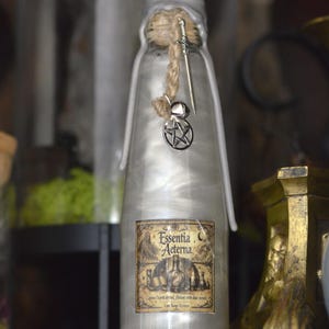 Peut inclure: Une bouteille en verre transparent remplie d'un liquide argenté scintillant, scellée avec de la cire blanche et de la ficelle. Une breloque épée et un pentagramme pendent au col. L'étiquette porte le texte Essentia Aeterna et une illustration.