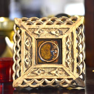 May include: A square, wooden frame with a carved, scalloped edge surrounds a ceramic tile. The tile features a crescent moon with a face in shades of brown and gold. The frame has a natural wood finish.