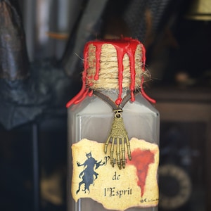 Puede incluir: Una botella de vidrio con una etiqueta de papel marrón que dice "Potion de l'Esprit". La botella está sellada con cera roja y tiene un amuleto de mano de esqueleto colgante.