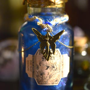 Op de afbeelding: Een blauwe glazen fles met een gouden dop en een feeënbedel. De fles heeft een etiket met de tekst "Fairy Honey".