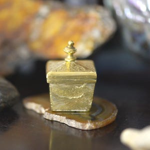Peut inclure: Petite boîte carrée dorée avec couvercle décoratif et fleuron. La boîte repose sur une tranche d'agate marron et beige. La boîte a une surface texturée et semble être en métal. L'arrière-plan est flou.