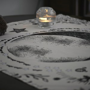 May include: A white altar cloth with a black crescent moon design, surrounded by a circle of small black hearts. There are two crystals, a star, and a lit candle on the cloth.