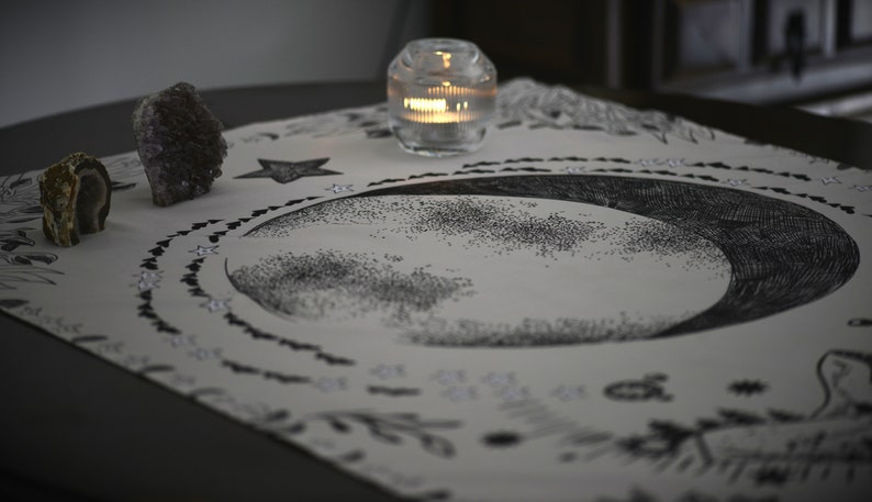 May include: A white altar cloth with a black crescent moon design, surrounded by a circle of small black hearts. There are two crystals, a star, and a lit candle on the cloth.