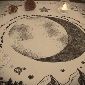May include: A beige cloth with a black and white crescent moon design. The moon is surrounded by a circle of small stars. There are two small crystals in the top left corner of the image.