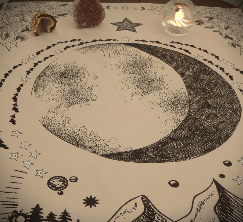 May include: A beige cloth with a black and white crescent moon design. The moon is surrounded by a circle of small stars. There are two small crystals in the top left corner of the image.