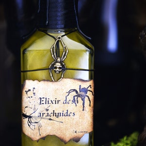 May include: A glass bottle with a black label that reads "Elixir des arachnides". The bottle is filled with a yellow liquid and has a black spider charm hanging from the neck.