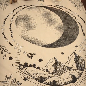 May include: A beige cloth with a black and white illustration of a crescent moon, stars, mountains, and trees. The cloth is decorated with a black border of stars and leaves.