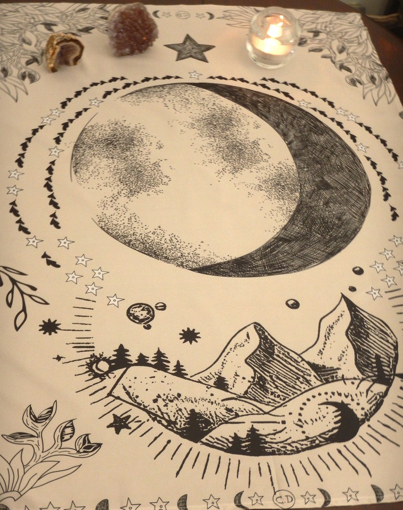 May include: A beige cloth with a black and white illustration of a crescent moon, stars, mountains, and trees. The cloth is decorated with a black border of stars and leaves.